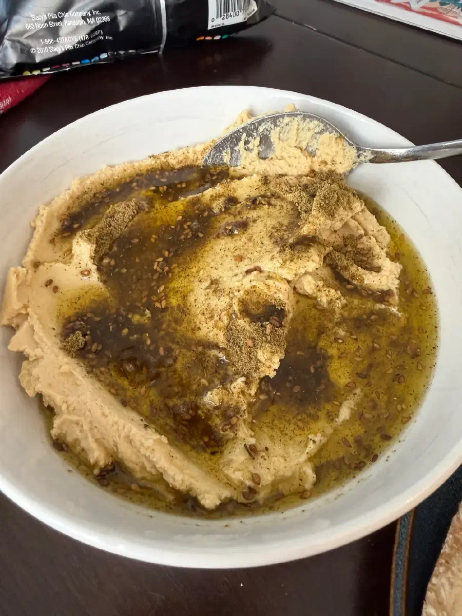 A white bowl of hummus topped with za'atar and a generous pool of olive oil, with a spoon resting in it