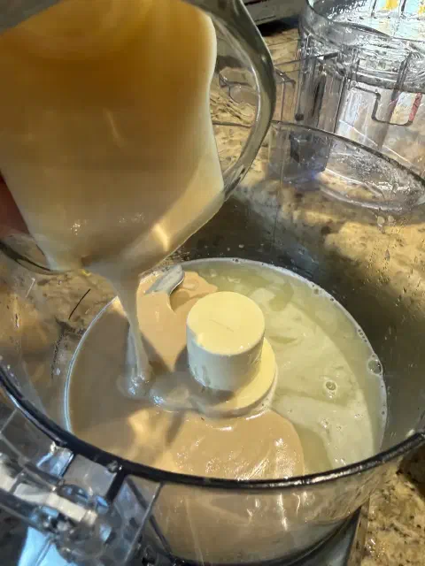 Tahini pouring from a measuring cup into the lemon juice in the food processor