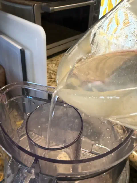 A thin stream of water pouring from a measuring cup into the food processor's feed tube
